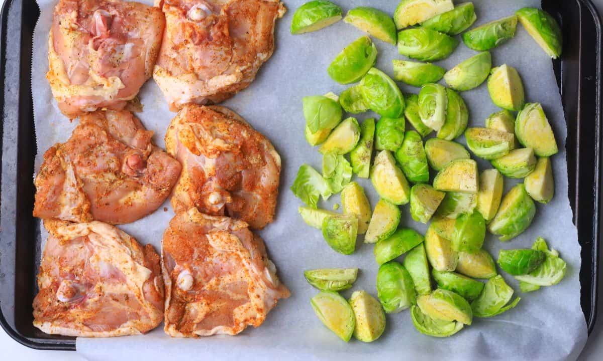 Raw chicken thighs with brussels sprouts on a parchment lined baking sheet.